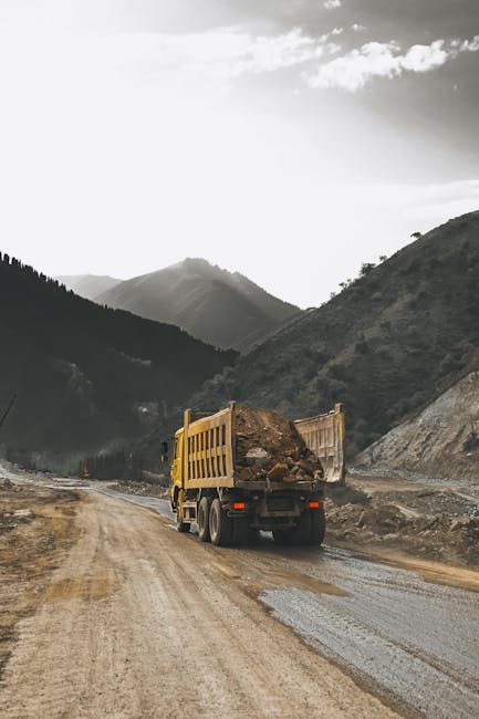 A yellow construction truck with a hydraulic lift bed is driving along a dirt road surrounded by mountainous terrain. The truck's bed is filled with large rocks and soil, indicating construction or land clearing activity. The road appears uneven and muddy, with patches of gravel visible. In the background, steep hills covered with dense greenery extend into the distance, under a partly cloudy sky. The scene conveys a remote, rugged environment, with no other vehicles or structures visible. Although the image is not directly related to house removals or moving services, it depicts heavy-duty transportation and logistical work that can be relevant to large-scale or construction-related relocations, aligning with themes of moving operations and transport logistics that [manwithvanwoodford.co.uk] might be involved with.