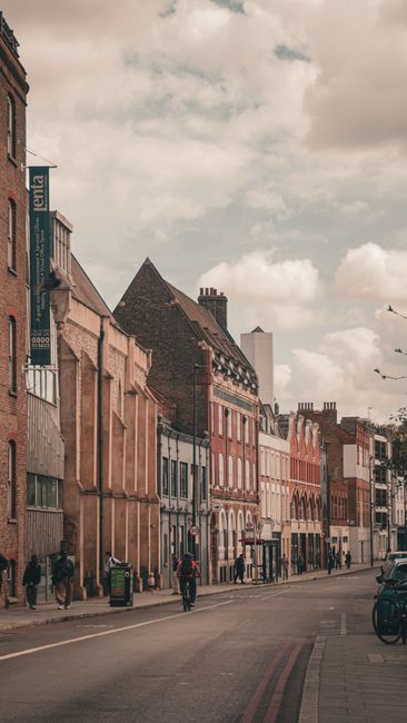 A street scene during daytime showing a row of multi-storey residential buildings with brick and concrete facades, some with bay windows and chimneys. In the foreground, a wide pavement accommodates s