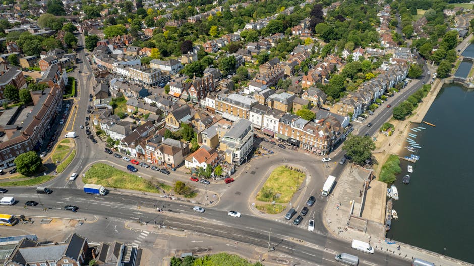 A street scene during daytime showing a row of multi-storey residential buildings with brick and concrete facades, some with bay windows and chimneys. In the foreground, a wide pavement accommodates s