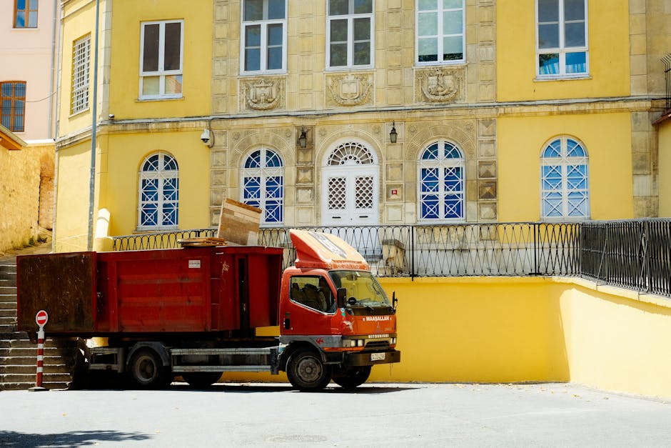A red removal truck parked on a paved area outside a yellow, multi-storey building with decorative stonework and arched windows. The truck's cargo area contains cardboard boxes and wooden furniture, indicating ongoing home relocation or furniture transport. The vehicle is positioned near a set of stairs and a yellow wall, with a metal railing visible along the upper level of the building. Bright daylight illuminates the scene, capturing the loading process associated with house moves, and the setting suggests an urban environment suitable for professional removals services by companies like Man with Van Woodford.