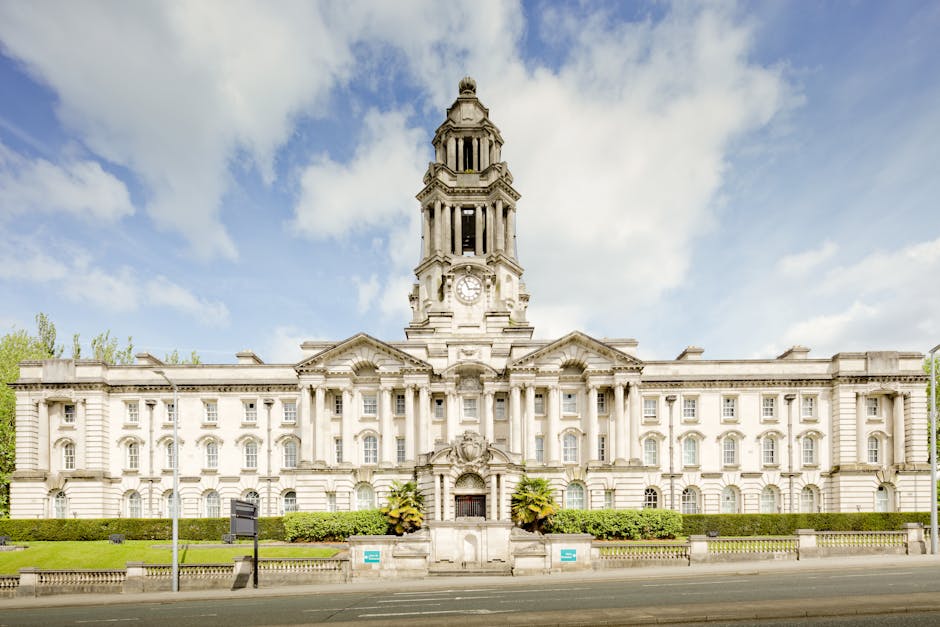 A grand, historic government building with a prominent central clock tower, constructed from light-colored stone and featuring ornate architectural details, including rounded windows and decorative columns. The building is set behind a low hedge and situated along a paved street. The sky above is partly cloudy with patches of blue. This image relates to house removals and logistics, reflecting the importance of proper handling of personal belongings during a home relocation process, adhering to regulations such as council fines for illegal removals, as discussed on the page for Man with Van Woodford.