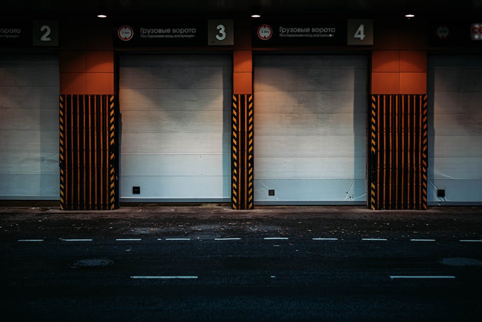 A street-level view of a commercial loading bay with three large, closed roller shutter doors numbered 2, 3, and 4, set in a modern building with white wall panels and red-orange vertical sections above each shutter. The loading bay area is framed by black and yellow striped protective barriers on either side of each door, emphasizing safety and designated loading zones. The pavement in front of the bays is dark asphalt with white road markings, and there are small patches of dirt or debris along the curb. Above the loading bays, there are signage boards with Russian text indicating truck gate and entrance information, suggesting this location is near a commercial or warehouse facility. This environment illustrates an organized space used for furniture transport and relocating household items, highlighting the logistics involved in home removals, as managed by companies like Man with Van Woodford, which specializes in packing and moving services.
