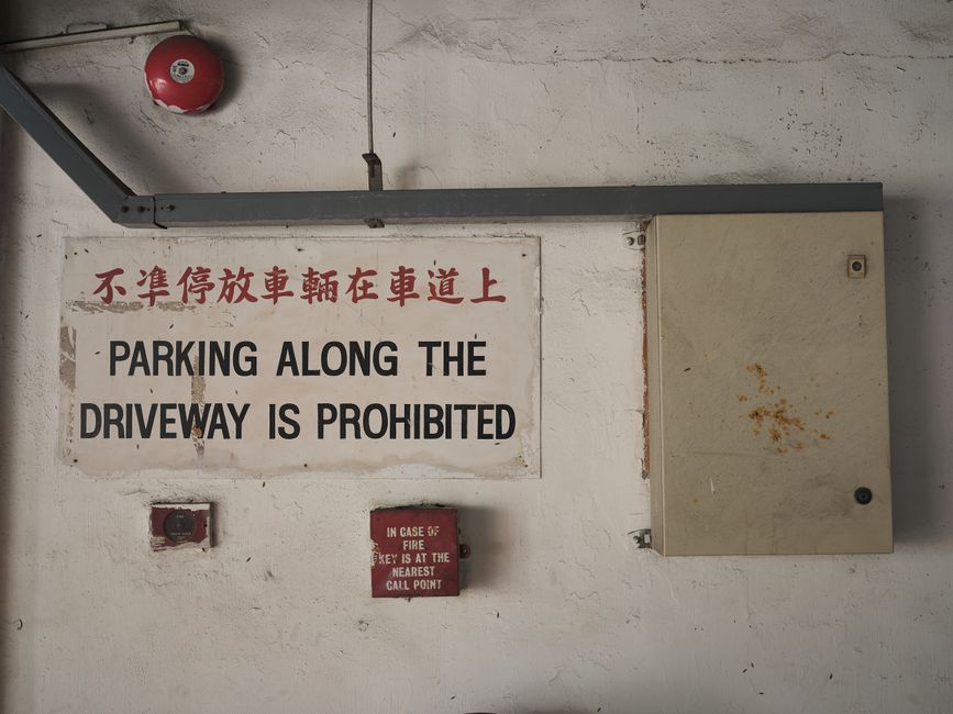 A white wall in a parking garage with a rectangular metal sign featuring Chinese characters and English text stating, 'PARKING ALONG THE DRIVEWAY IS PROHIBITED.' Below the sign, there are two small red emergency devices; one indicates a fire alarm with the instruction to call the nearest fire station in case of activation. Above the sign, a metal conduit pipe runs horizontally, and a large beige electrical panel with rust spots is mounted on the right side of the wall. The setting is a utilitarian indoor space, likely used for house removals or furniture transport, where clear signage about parking restrictions is important for planning and access during moving processes. The lighting is neutral, highlighting the functional environment typical of a building’s parking or delivery area, relevant to professional removals services like [COMPANY_NAME].