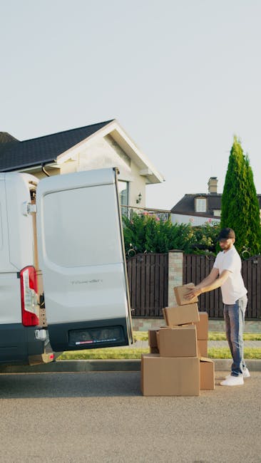 Two professional movers from Man with Van Woodford are loading a large, dark green upholstered item wrapped in protective fabric into the rear of a white cargo van parked on a street near Woodford Sta