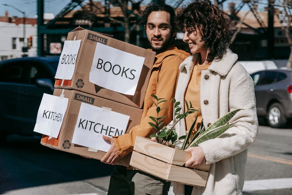 A young man and woman are outside in a street, engaged in a home relocation or packing process. The man is carrying two cardboard boxes labeled 'BOOKS' and 'KITCHEN' with white paper and black text, i