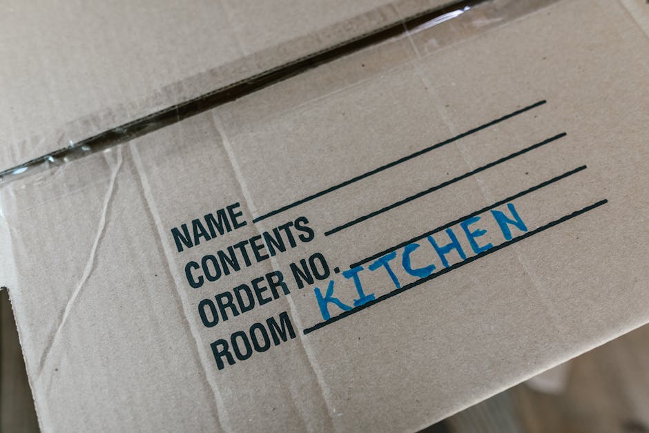 A young man and woman are outside in a street, engaged in a home relocation or packing process. The man is carrying two cardboard boxes labeled 'BOOKS' and 'KITCHEN' with white paper and black text, i