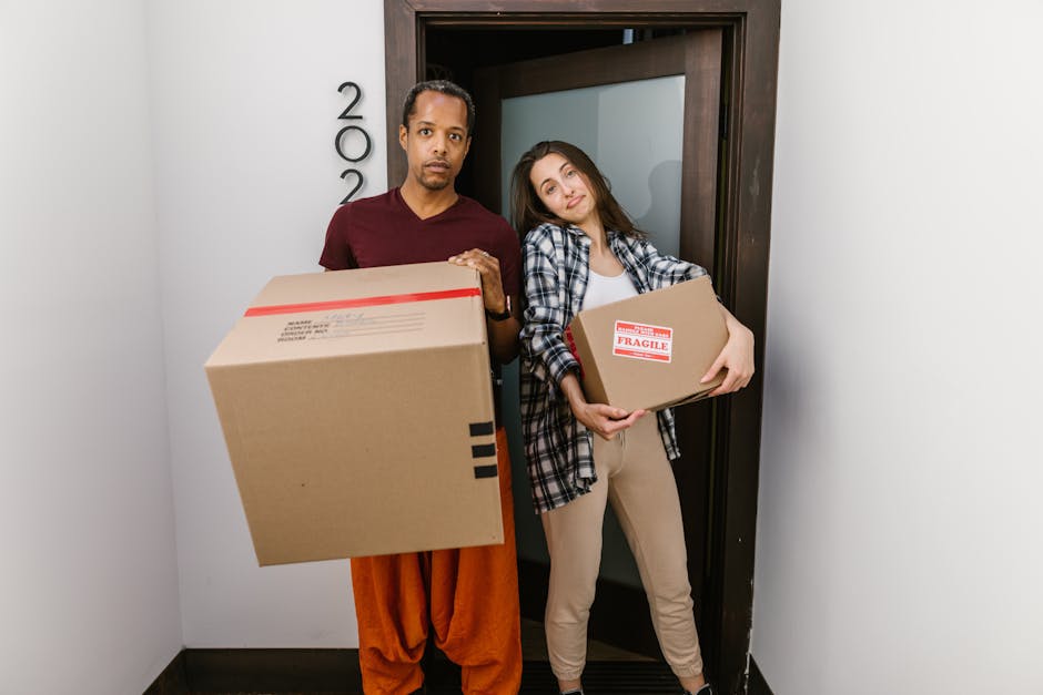 A man and a woman stand inside an entryway near a door, each holding a cardboard box for home relocation. The man, on the left, wears a maroon t-shirt and orange pants, with a neutral expression, holding a large box with a red tape seal. The woman, on the right, wears a white top and plaid shirt, with beige pants, and is leaning her head slightly towards the man while holding a smaller box marked 'Fragile.' Behind them, the door frame is visible with a partially opened door leading to another room, and the surroundings reveal a clean, organized indoor environment with smooth grey flooring and white walls, indicative of being inside a residential property ready for packing and moving. The boxes appear to be made of sturdy cardboard, suitable for transporting household items during a house removal process, with no visible packaging materials or equipment in the scene.