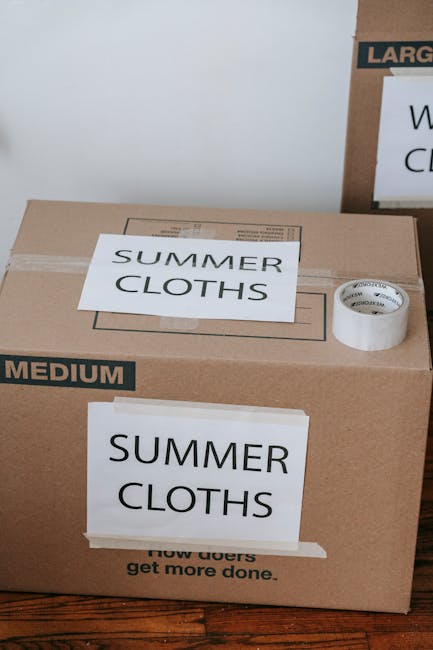 A large cardboard box labeled 'SUMMER CLOTHS' and 'MEDIUM' is situated on a wooden floor inside a property, ready for home relocation or packing as part of a moving process. The box is sealed with packing tape, and a roll of clear adhesive tape is placed on top. Beside it, there is another cardboard box partially visible, indicating an organized packing setup typical of furniture transport and household moves carried out by Man with Van Woodford. The environment appears well-lit, with neutral background walls. The boxes are likely prepared for loading into the van during the furniture transport phase, supporting efficient packing tips for flats on Chingford Road, Woodford, and the ongoing moving logistics involved in house removals.