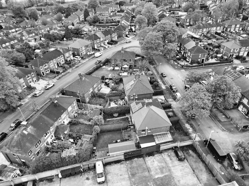 An aerial view of a residential neighbourhood showing several houses, gardens, and parked cars along the street. In the foreground, a house with a sloped roof is visible, with a fenced garden area adjacent to it. There are various cardboard boxes, wrapped furniture, and moving blankets visible within the property, indicating packing materials for a home relocation. A van is parked nearby, and part of the loading process appears to be taking place with furniture being moved from the house onto the vehicle. The surroundings include additional houses and trees, with clear road access and pavement. The image exemplifies typical scenes involved in furniture transport and packing during a professional removals service, such as those provided by Man with Van Woodford, adhering to proper moving and logistical procedures based on the local Redbridge Council permit rules.