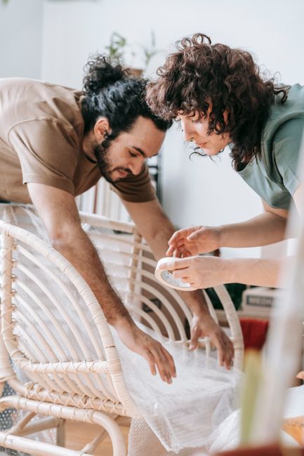 Two individuals are engaged in packing or preparing furniture for a home relocation inside a well-lit room. One person, with curly dark hair and wearing a beige shirt, is carefully wrapping a piece of