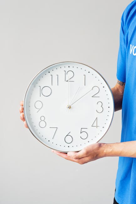 A person wearing a blue shirt is holding a large white wall clock with a grey frame, displaying the time as approximately 12:07. The individual is grasping the clock with both hands, one hand supporting the bottom and the other holding the side. The clock has a minimalistic design with simple grey numbers and hands, and is positioned against a plain light grey background. The scene suggests a move or packing situation, consistent with house removals or home relocation services provided by Man with Van Woodford. The image captures the process of handling household items, possibly during a packing, loading, or moving preparation phase, which aligns with the context of urgent removals and furniture transport.