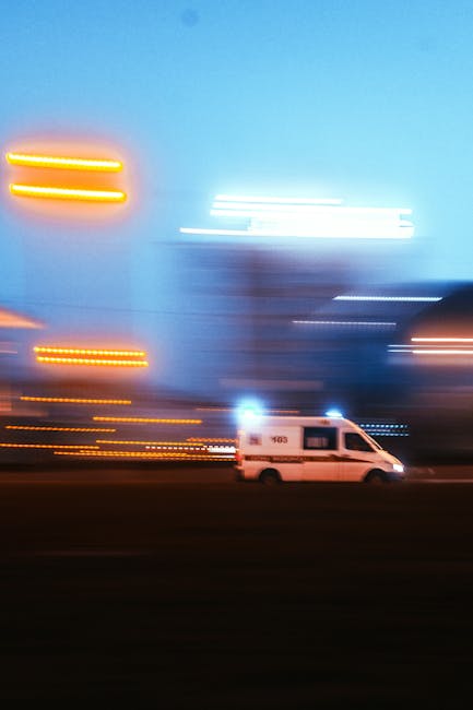 A white van with the number 193 on its side is parked on a street at dusk, with illuminated cityscape buildings in the background. The photo captures a motion blur effect, showing streaks of light from streetlights and building signs, indicating a night-time urban environment. The van appears to be positioned for potential furniture transport or part of a house removal process. Based on the context of house removals and logistic services, this scene suggests the vehicle may be involved in a home relocation operation managed by Man with Van Woodford, serving clients needing urgent or efficient moving and packing services within Woodford.
