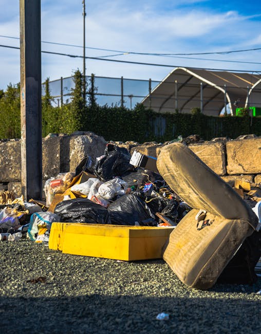 A pile of mixed bulky waste including several black garbage bags, a yellow plastic storage box, a large discarded car tire, and various cardboard and plastic packaging materials situated on a gravel surface beside a stone wall. The waste appears to be collected for disposal or removal, possibly related to home relocation or clearance services. In the background, a metal fence, a utility pole, and some trees are visible beneath a partly cloudy sky. The scene is illuminated by natural daylight, highlighting the different textures and materials of the waste items. This setting likely depicts an outdoor area prepared for waste collection or clearance as part of a moving or rubbish disposal process, with a focus on furniture or household item removal at a property in Woodford, aligning with services offered by Man with Van Woodford.