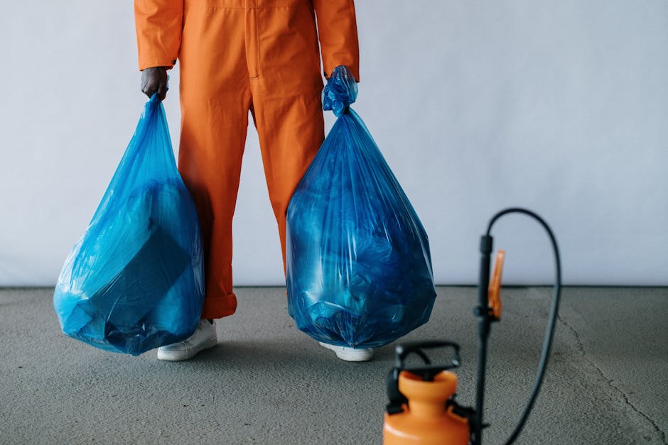 A person wearing orange work overalls stands inside a room with plain, light-colored walls and a grey carpeted floor, holding two large blue plastic bags filled with waste or belongings, one in each hand. The individual’s face is not visible, as the image is cropped at the waist. In front of them, on the left side of the image, an orange and black handheld device or sprayer is seen, likely part of cleaning or waste disposal equipment. The setting appears to be part of a house removal process, where packing, packing materials, or waste are being managed. The scene emphasizes the task of clearing bulky waste during a home relocation, aligning with services offered by [COMPANY_NAME], such as removals and waste disposal, as part of a professional moving or clearance process involved in house removals in Woodford.